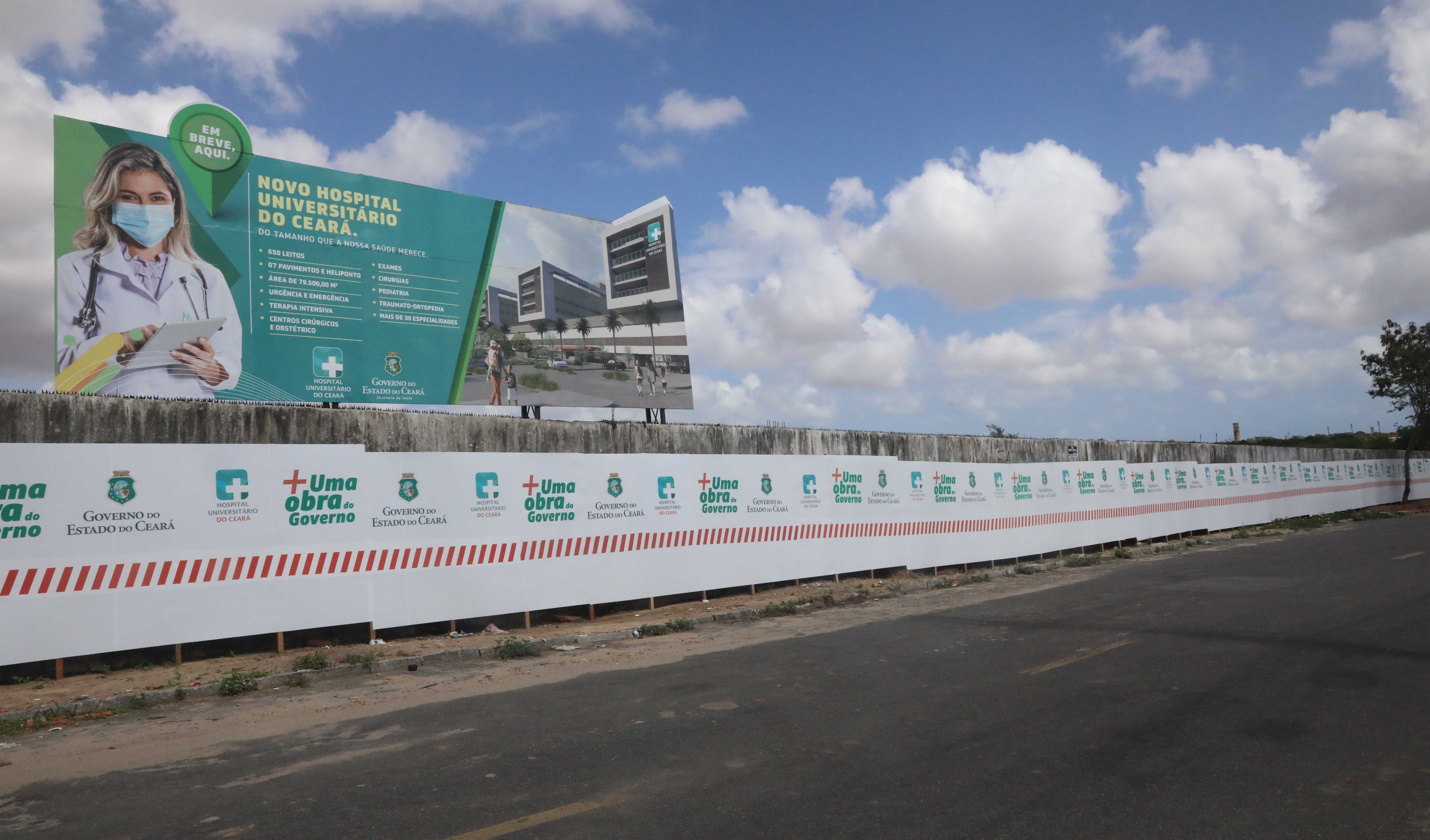 grande muro com várias marcas do Governo do Estado em outdoor acima com nome do hospital e foto de moça vestida de médica